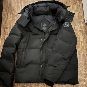 Svart dunjacka från Canada Goose M - Säljer en svart dunjacka från Canada Goose i storlek M. Jackan har en klassisk puffer-design med huva och avtagbar pälsdetalj. Märket syns tydligt på ärmen och insidan. Jackan har både dragkedja och knappar framtill för extra skydd mot kyla. 