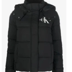 Svart pufferjacka från Calvin Klein - Svart pufferjacka från Calvin Klein Jeans med stor logga på bröstet och ärmen. Jackan har huva, dragkedja och knappar framtill. Fylld med syntetmaterial för extra värme och har en clean, boxig passform som är perfekt för kalla dagar.