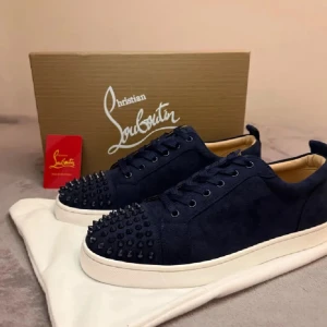 Mörkblå sneakers från Christian Louboutin - Snygga mörkblå sneakers från Christian Louboutin med mockaöverdel och coola svarta nitar på tån. Skorna har snörning, vit platt sula och den ikoniska röda undersidan. Perfekt för dig som vill sticka ut med exklusiv stil.