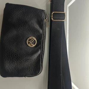 Svart axelväska med guldiga detaljer - Svart axelväska i skinn från märket NYPD med avtagbar kedja❤️i ena facket förekommer det lite rosa färg som inte går bort men ör verkligen inte synligt❤️