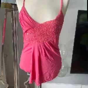 Glittrig rosa linnetopp med paljetter - Säljer en snygg rosa linnetopp med smala axelband och glittriga paljetter över bysten. Toppen har en omlott-look framtill och är figurnära med ett mjukt fall nedtill. Perfekt för dig som vill sticka ut med lite extra bling och färg.