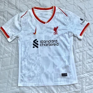 Liverpool Salah bortatröja Nike vit - Liverpool FC bortatröja i vitt med röda och gula detaljer från Nike. Tryck med M. Salah och nummer 11 på ryggen. Tröjan har korta ärmar, V-ringning och diskret mönster i tyget. Materialet är lätt och andas, perfekt för fotboll eller supporterliv. Passar folk mellan 152-160cm