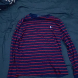 Randig långärmad tröja Polo Ralph Lauren - Snygg långärmad tröja från Polo Ralph Lauren i marinblått med röda ränder. Klassisk rund hals och diskret logga på bröstet. Perfekt för dig som gillar stilrena och sportiga vibes. Materialet är mjuk bomull som känns skönt mot huden.