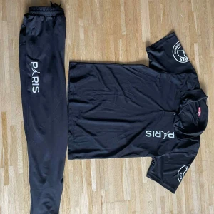 Svart PSG x Jordan pikétröja - Svart pikétröja från Jordan x PSG med vit Paris-logga på bröstet och ärmen. Tröjan har klassisk krage, korta ärmar och knappslå. Materialet är mjukt och lätt polyester, perfekt för sport eller chill. Snyggt tryck på både fram- och baksida.