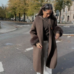 Sydney lång brun kappa från Femenina - Supersnygg lång brun kappa från Femenina of Sweden med breda ärmar och fluffiga pälsdetaljer vid ärmsluten. Kappan har omlottmodell med bälte i midjan och en elegant.             Storlek S. Passar tyvärr inte mig. Prislapp finns kvar