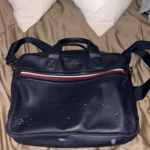 Mörkblå datorväska Tommy Hilfiger - Snygg mörkblå datorväska från Tommy Hilfiger med axelrem och handtag. Väskan har en dragkedja framtill med röd, vit och blå detalj samt diskret logga. Perfekt för att bära laptop och viktiga grejer till plugget eller jobbet. Den har små  märken/förg förändring.