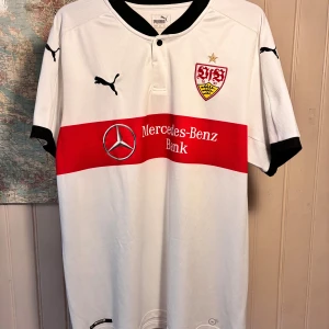 VfB Stuttgart Puma 2017/18 home shirt, size XL - Säljer en officiell VfB Stuttgart fotbollströja från Puma i storlek XL. Tröjan är vit med en bred röd horisontell rand över bröstet, svart krage och svarta detaljer vid ärmslut. Materialet är lätt och andas, perfekt för match eller träning. Klubbmärke och Puma-loggor på bröstet.