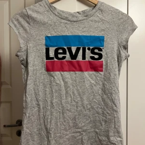 Grå Levi's t-shirt med logga - Snygg grå t-shirt från Levi's med klassisk logga i blått, svart och rött framtill. T-shirten är kortärmad och har en rund halsringning. Perfekt för en avslappnad och cool stil.