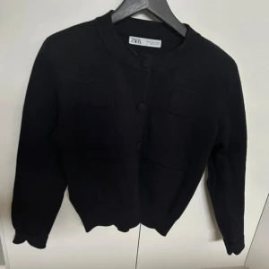 Svart kofta från Zara med knappar - Stilren svart kofta från Zara med rund halsringning och dekorativa knappar framtill. Koftan har två bröstfickor och två fickor nertill, samt långa ärmar. Perfekt att slänga på sig för en clean och enkel look.