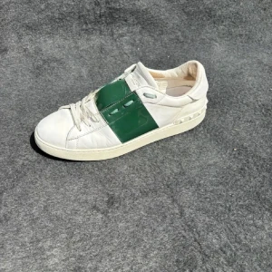 Valentino Open sneakers vit/grön - Snygga Valentino Open sneakers i vitt skinn med en grön lackad panel över mitten och coola nitar på hälen. Rund tå, platt sula och klassisk snörning. Perfekt för dig som gillar exklusiva och trendiga sneakers med unik design.