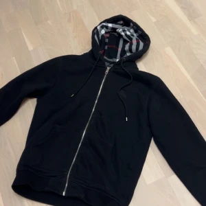 Svart Burberry hoodie med dragkedja - Svart hoodie från Burberry med klassiskt rutigt foder i huvan i grått, vitt och rött. Tröjan har hel dragkedja, snörning i huvan och två fickor framtill. Tillverkad i mjuk bomull för en skön känsla. Perfekt för dig som vill ha en stilren och lyxig look.