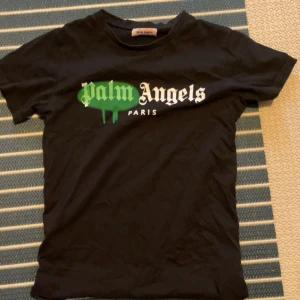 Svart Palm Angels t-shirt med tryck - Svart t-shirt från Palm Angels med grönt och vitt tryck på bröstet där det står 'Palm Angels Paris'. Klassisk passform med rund hals och korta ärmar. Materialet är mjuk bomull, perfekt för en avslappnad streetwear-look.
