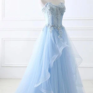 Ljusblå långklänning med spetsdetaljer - Drömmig ljusblå långklänning med axelbandslös överdel, dekorerad med vackra spetsapplikationer och blommiga detaljer. Kjolen är i flera lager av tunn tyll som ger en svävande känsla. Perfekt för bal eller speciella tillfällen.