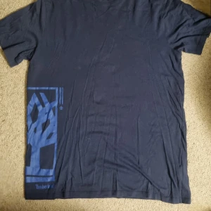 Mörkblå Timberland t-shirt i bomull - Snygg mörkblå t-shirt från Timberland i 100% ekologisk bomull. T-shirten har korta ärmar och ett coolt blått tryck med Timberland-loggan på sidan. Perfekt för en avslappnad och stilren look.