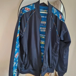 Adidas blå bomberjacka med mönster - Säljer en blå bomberjacka från Adidas med färgglatt retroinspirerat Adidas-mönster på insidan och längs ärmarna. Jackan har dragkedja, ribbade muddar och klassisk Adidas-logga på bröstet. Perfekt för dig som vill sticka ut med en unik streetstyle.