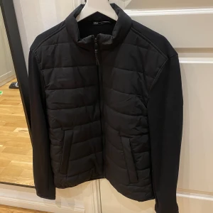 Svart quiltad pufferjacka från Zara - Svart pufferjacka från Zara i storlek L. Jackan har quiltad framsida, släta ärmar och hög krage. Stängs med dragkedja och har två snedställda fickor framtill. Perfekt för kyliga dagar och enkel att matcha med det mesta.