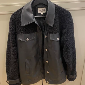 Svart teddyjacka från NA-KD, stl 34 - Snygg svart teddyjacka från NA-KD i storlek 34. Jackan har en cool mix av teddyfleece på ärmar och axlar samt släta partier i syntetmaterial. Stora fickor framtill, silverfärgade knappar och bred krage ger en trendig look. Små defekter på kragen (se bild) men syns inte vid användning 