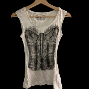Vit linne-topp med korsettprint Guess - Cool vit ärmlös linne-topp från Guess med ett svart tryck som föreställer en korsett framtill oc baktill. Rund halsringning och mjukt bomullsmaterial gör den både bekväm och snygg. 