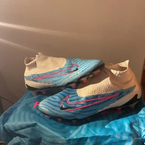 Nike Phantom GX Elite fotbollsskor blå/vit - Helt sprillans nya Nike Phantom GX Elite fotbollsskor i blått och vitt med rosa detaljer och svart Nike-logga. Som är köpta från unisport. Skorna har en strumpliknande passform utan snörning, mönstrad ovandel och platt sula med dobbar för gräsplan. Tillverkade i syntetmaterial för maximal känsla och kontroll. 