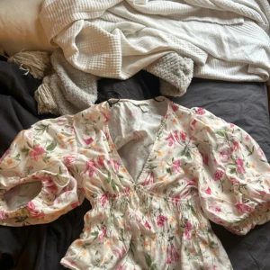 Blommig kortklänning från H&M XS - Supersöt blus från H&M i storlek XS med blommigt mönster i rosa, grönt och gult på vit botten. Klänningen har puffiga korta ärmar, v-ringning och smock i midjan som ger en snygg passform. Perfekt för vår och sommar!