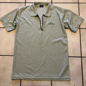 Ljusgrå randig pikétröja från Okey - Säljer en ljusgrå pikétröja från Okey med diskreta vertikala ränder och halv dragkedja framtill. Tröjan har korta ärmar och klassisk krage, perfekt för en chill och sportig look. Broderad logga på bröstet.