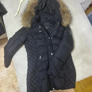 Svart dunjacka Rockandblue med päls - Svart lång dunjacka från Rockandblue med snygg quiltad design och avtagbar huva med päls.Jackan har flera dragkedjefickor, emblem på ärmen och markerad midja för en schysst siluett. Perfekt för kalla dagar.