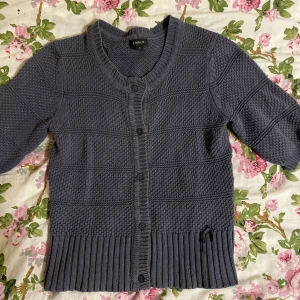 Grå stickad kofta från Lindex - Mysig grå stickad kofta från Lindex med rund hals och knappar framtill. Koftan har ett snyggt strukturerat mönster, ribbad nederkant och en liten rosett-detalj vid höften. Ärmarna är lite kortare, perfekt för lager-på-lager. Passar dig som gillar klassisk stil med en twist.