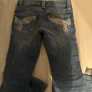 Blå bootcut jeans med broderade fickor - Snygga blå jeans från Gina Young med bootcut och lågmidjad passform och slitna detaljer framtill. Baktill har de två fickor med silverfärgat broderi i vingar, vilket ger en unik look.