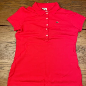 Röd Lacoste piké t-shirt - Snygg röd piké t-shirt från Lacoste med klassisk krage och korta ärmar. Tröjan har knappar framtill och den ikoniska gröna krokodilloggan broderad på bröstet. Tillverkad i mjuk bomull som andas och passar perfekt till en chill stil.