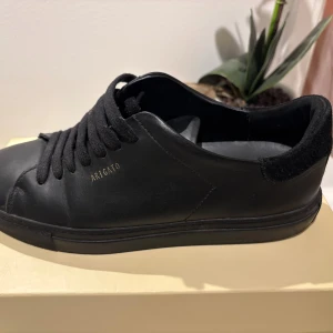 Svarta sneakers från Axel Arigato - Stilrena svarta sneakers från Axel Arigato i skinn med diskret guldfärgad logga på sidan. Skorna har svart mockadetalj vid hälen, svarta snören och en klassisk låg profil. Perfekta för dig som gillar minimalistisk och clean design.