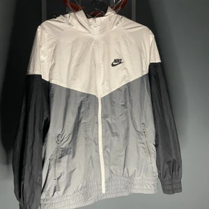 Nike vindjacka i svart och vitt XL - Nike vindjacka med huva i svart, grått och vitt. Jackan har dragkedja framtill, två sidofickor med dragkedja och elastiska muddar. Lätt och sportig design med klassisk Nike-logga på bröstet. Perfekt för blåsiga dagar och enkel att packa ner.