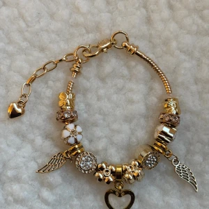 Guldigt Pandora armband med berlocker - Unikt Pandora armband i guldton med flera berlocker, bland annat blommor, hjärta, änglavingar och glittriga detaljer. Vissa berlocker har vit emalj och strass. Perfekt för dig som gillar personliga smycken med mycket detaljer och charm.