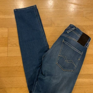 Replay Anbass blå jeans, storlek 29 - Snygga blå Replay Anbass jeans med klassisk femficksdesign och coola slitningar framtill. Jeansen har raka ben, normal passform och tydliga sömmar på bakfickorna. Perfekta för dig som gillar en stilren och modern look.