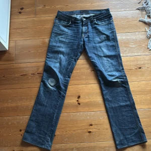 Blå jeans från Nudie Jeans W34 L36 - Säljer ett par klassiska blå jeans från Nudie Jeans med snygga slitningar och kontrastsömmar. Modellen har raka ben, fem fickor och cool broderad detalj på bakfickorna. Passar dig som gillar en avslappnad och tidlös jeansstil.