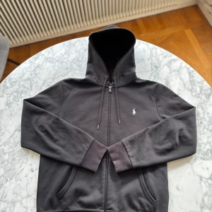 Svart hoodie från Polo Ralph Lauren - Svart hoodie från Polo Ralph Lauren med klassisk logga broderad på bröstet. Den är använd rätt så många gånger men är i oväntat bra skick. Köpt för 3000 och är i storleken S. Perfekt om du gillar hoddies som sitter slim.