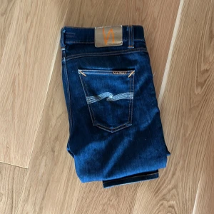 Mörkblå jeans från Nudie Jeans - Snygga mörkblå jeans från Nudie Jeans med klassisk femficksdesign och kontrastsömmar. Jeansen är tillverkade av ekologisk bomull och har en rak passform med smal siluett. Bakfickorna har Nudie Jeans signaturbroderi iljusblått