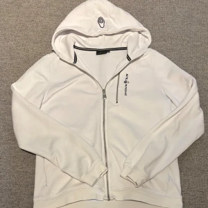Vit Sail Racing Zip Hood - Vit Zip hoodie från Sail Racing med dragkedja och huva. Tröjan har långärmade ärmar, ribbade muddar och ett broderat Sail Racing-tryck på bröstet. Materialet är mjukt och känns som bomull, perfekt för chill dagar. Snygg detalj med logga på huvan.