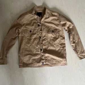 Beige overshirt från 157, storlek S - Snygg beige overshirt från 157 i storlek S. Jackan har två bröstfickor med knappar, klassisk krage och knäppning framtill. Tillverkad i bomull, perfekt för lager-på-lager och chill stil. Passar dig som gillar enkel och clean look.