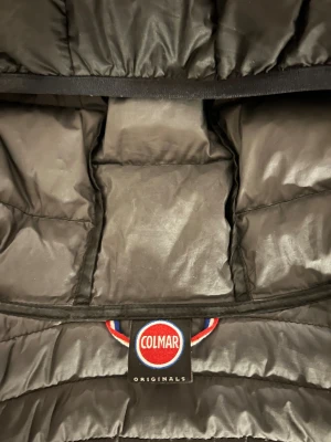 Svart dunjacka med huva från Colmar - Snygg svart dunjacka från Colmar Originals med huva och två dragkedjeförsedda fickor. Jackan är quiltad och har en smidig passform, perfekt för kyliga dagar. Klassisk Colmar-logga på ärmen och insidan. Långärmad och lätt att bära.