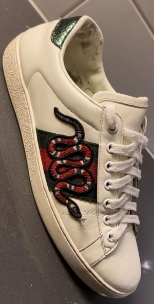Gucci Ace sneakers med orm - Gucci Ace sneakers i vitt skinn med klassiska gröna och röda ränder på sidan. Ena skon har en broderad orm i rött, svart och vitt. Baksidan har grön metallic-detalj med ormmönster. Snygga och ikoniska sneakers med snörning och platt sula.