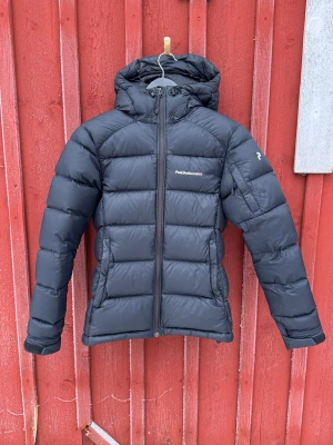 Svart dunjacka Peak Performance Frost Down XS - Säljer en svart dunjacka från Peak Performance i storlek XS. Jackan har huva, dragkedja framtill och klassisk pufferdesign med broderad logga på bröstet och ärmen. Perfekt för kalla dagar och riktigt snygg streetstil. Jackan är i nyskick och sparsamt använd.