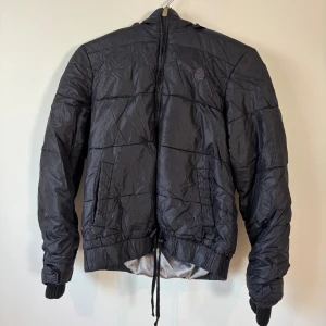 Vero Moda pufferjacka  - M storlek - Svart pufferjacka från Vero Moda med broderad logga på bröstet. Jackan har huva, dragkedja framtill, ribbade muddar och snörning i nederkant. 