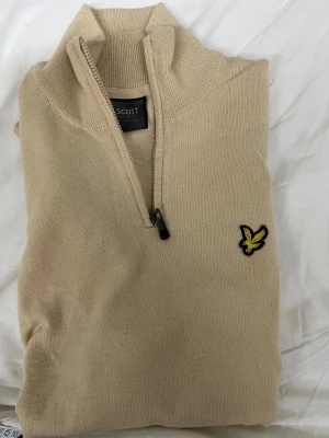 Beige half zip stickad tröja Lyle & Scott - Snygg beige stickad tröja från Lyle & Scott med half zip och klassisk gul fågel-logga på bröstet. Tröjan har hög krage och är tillverkad i mjuk bomull, perfekt för lager-på-lager under hösten och vintern.