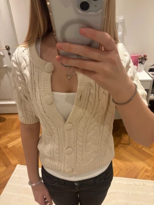 Beige stickad kofta från hm - Säljer en beige stickad kofta med korta ärmar och stora matchande knappar framtill. Koftan har ett snyggt kabelstickat mönster och ribbade kanter vid midja och ärmslut.