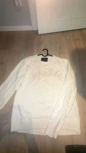 Vit stickad tröja från Morris - Vit stickad tröja från Morris med broderad logga i stora bokstäver över bröstet. Rund halsringning och ribbade muddar vid ärmslut och nederkant. Perfekt för dig som gillar klassisk och clean stil.finns en liten fläck på armen men den syns inte tydligt 