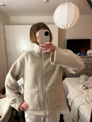 Beige teddyjacka med dragkedja S - Mysig beige teddyjacka i storlek S med hög krage och hel dragkedja framtill. Jackan har två stora fickor och är tillverkad i fluffigt fleece-material som håller dig varm under kyliga dagar. Perfekt för lager-på-lager och chill vibes. Har en liten fläck på fickan 