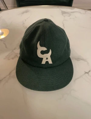 Mörkgrön keps från Chromakopia - Snygg mörkgrön keps med vit broderad CA-logga framtill och Chromakopil broderat baktill. Justerbar rem i brunt läder baktill. Kepsen har klassisk form och är tillverkad i ull för en cool och unik look.