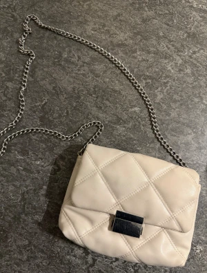 Beige quiltad axelväska med kedja - Snygg beige axelväska med quiltad design och silverfärgad kedja som axelrem. Väskan har ett stilrent metallspänne framtill och ett innerfack med dragkedja. Perfekt storlek för det viktigaste och ger en clean vibe till din outfit.