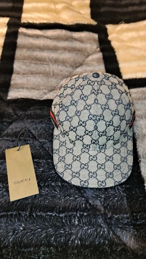 Gucci keps med GG-mönster och röd-blå detalj - Snygg keps från Gucci i beige och marinblått med klassiskt GG-monogram över hela kepsen. Kepsen har justerbar rem bak i blått skinn och röda/blå ränder på sidorna. Tillverkad i bomullscanvas med exklusiv känsla. Perfekt för dig som vill sticka ut med lyxig streetstyle. Helt ny oanvänd med etikett på
