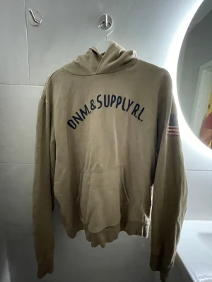 Beige hoodie från Ralph Lauren - Säljer en beige hoodie från Denim & Supply Ralph Lauren med tryck på bröstet och en amerikansk flagga patch på ärmen. Tröjan har huva, känguruficka och långa ärmar. Perfekt för chill dagar och har en avslappnad vibe.